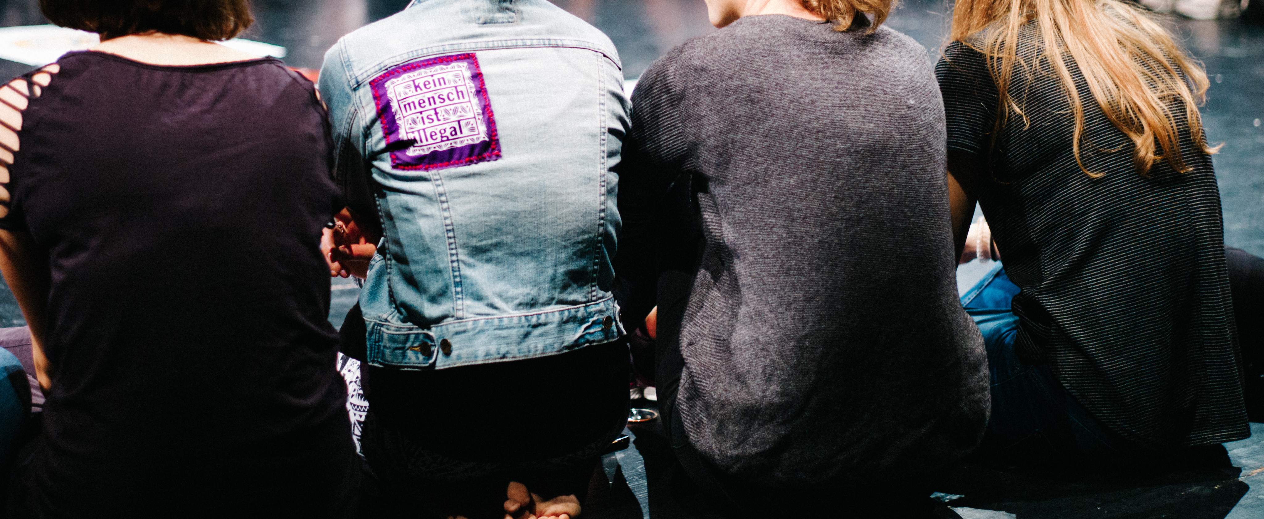 4 junge Menschen wenden uns den Rücken zu während Sie nebeneinander am Boden sitzen. Die Rückseite einer Jacke trägt einen Aufnäher mit der Aufschrift: "Kein Mensch ist illegal!".